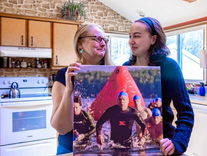 Maia Bradley and her mother, Miiko, hold up a photo of Mike Bradley in one of his triathlon photos in the family kitchen on Saturday, March 30. 2024.